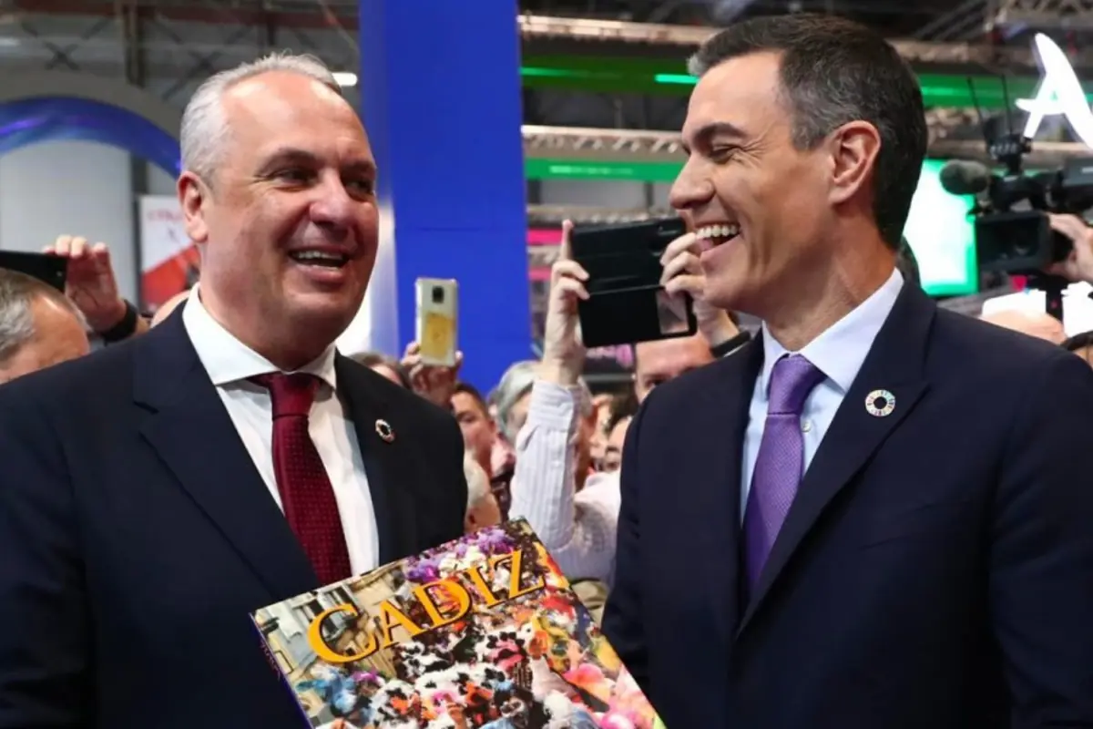 Dos hombres de traje sonríen mientras uno de ellos sostiene un libro con la portada que dice Cádiz en un evento con varias personas y cámaras al fondo