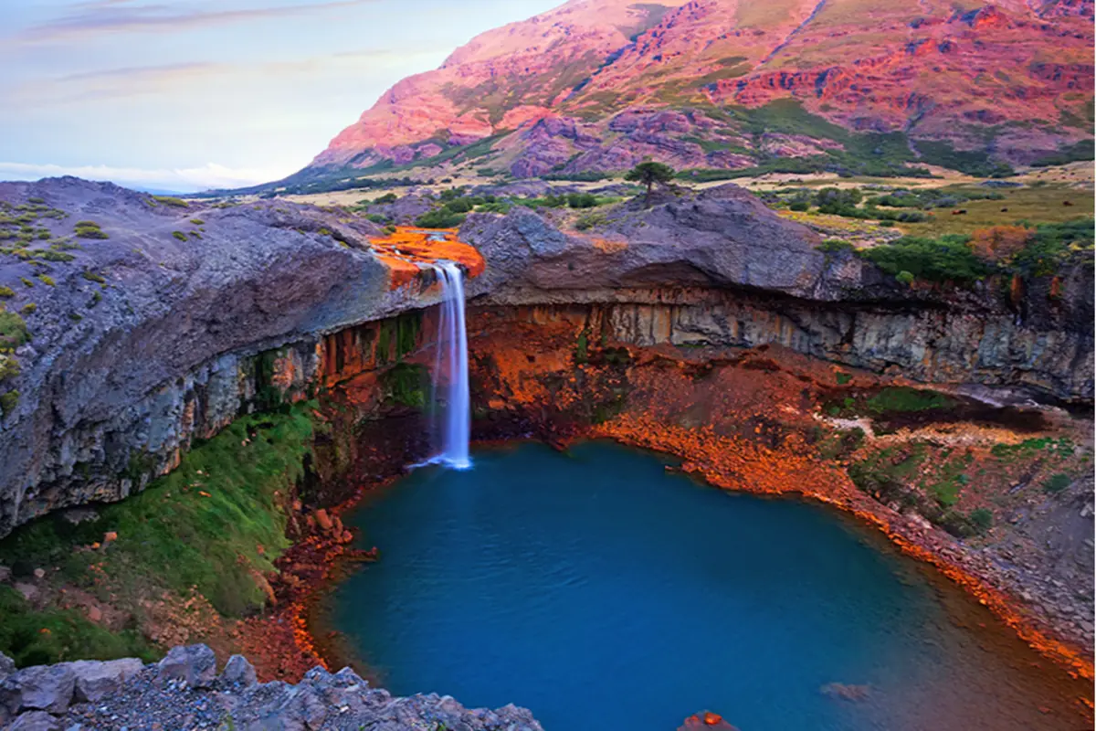 Cascada cayendo en una poza de agua azul rodeada de formaciones rocosas y montañas al atardecer