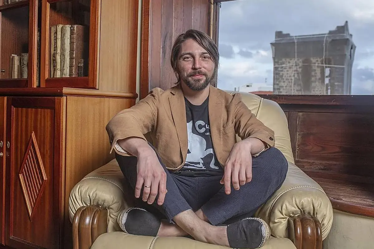 Hombre sentado en un sillón beige con las piernas cruzadas, vistiendo un saco marrón y camiseta negra, en una sala con librero de madera y una ventana al fondo