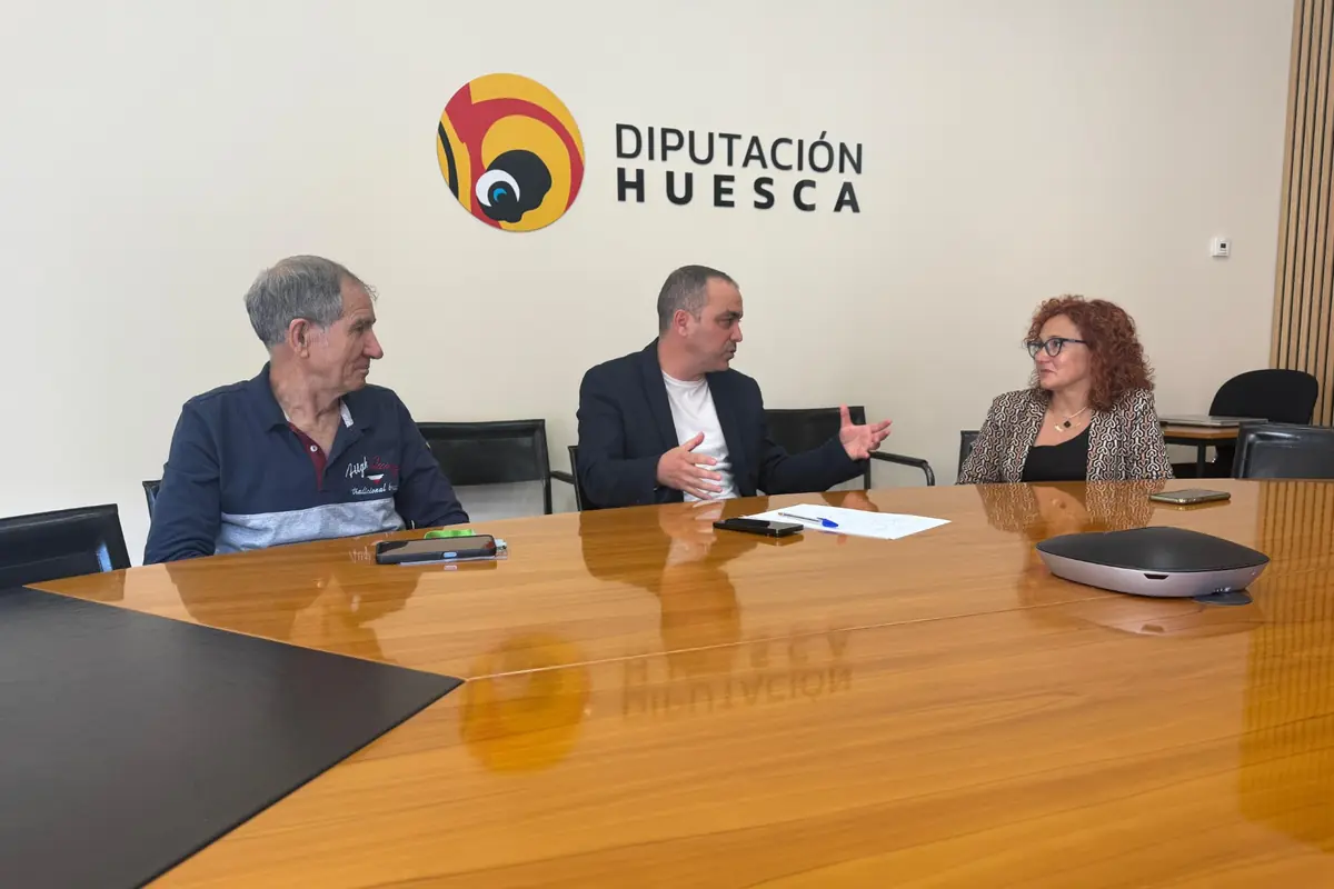 Tres personas conversan sentadas alrededor de una mesa de reuniones en una sala con el logo de Diputación Huesca en la pared.