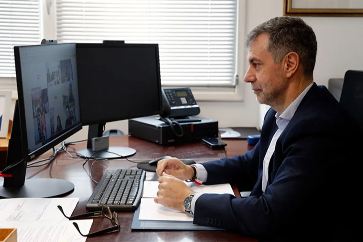 Hombre de mediana edad con barba sentado en un escritorio trabajando en una videollamada frente a dos monitores en una oficina iluminada por luz natural Hombre de mediana edad con barba sentado en un escritorio trabajando en una videollamada frente a dos monitores en una oficina iluminada por luz natural