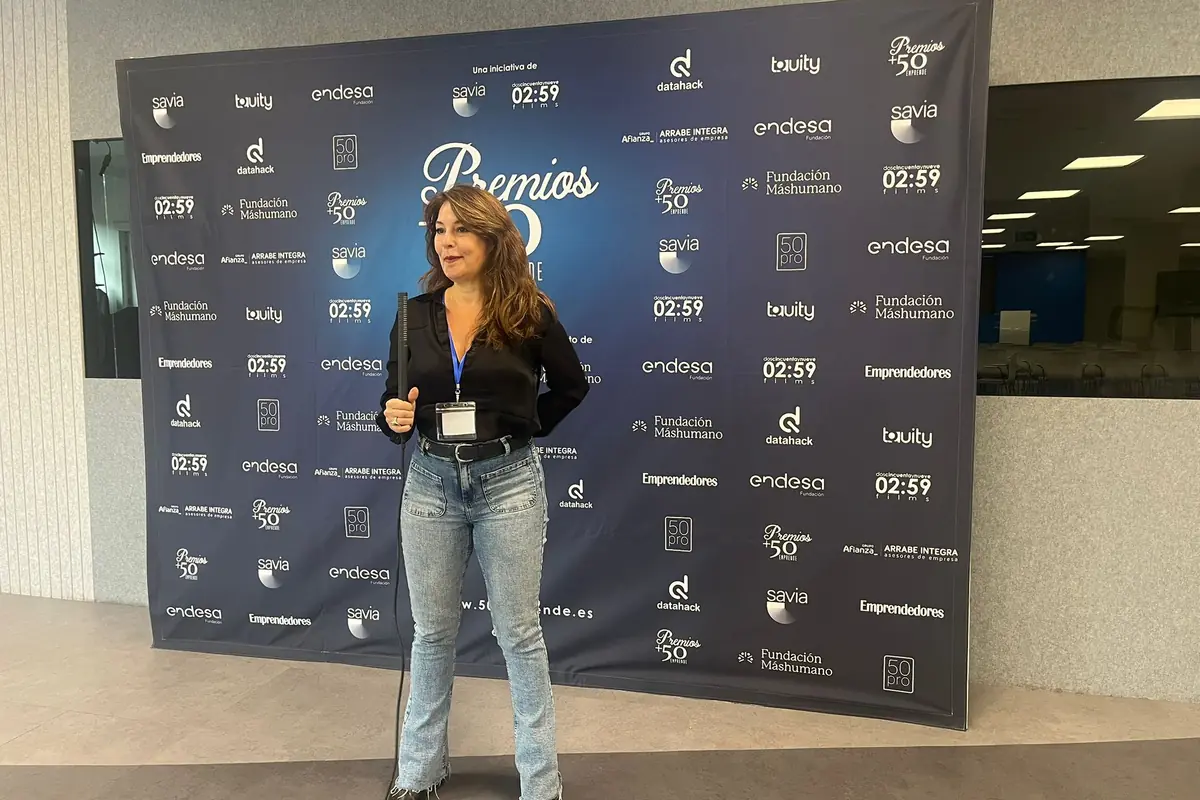Mujer de cabello largo y castaño vestida con blusa negra y jeans posando frente a un fondo azul con logotipos de patrocinadores en un evento de premios