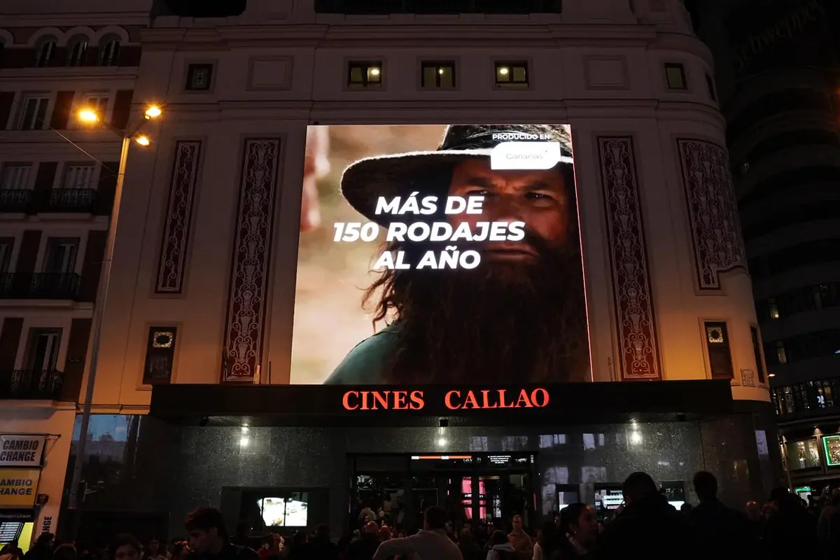 Fachada de Cines Callao iluminada de noche con una pantalla gigante que muestra la imagen de un hombre con barba y sombrero y el texto más de 150 rodajes al año mientras varias personas caminan por la plaza