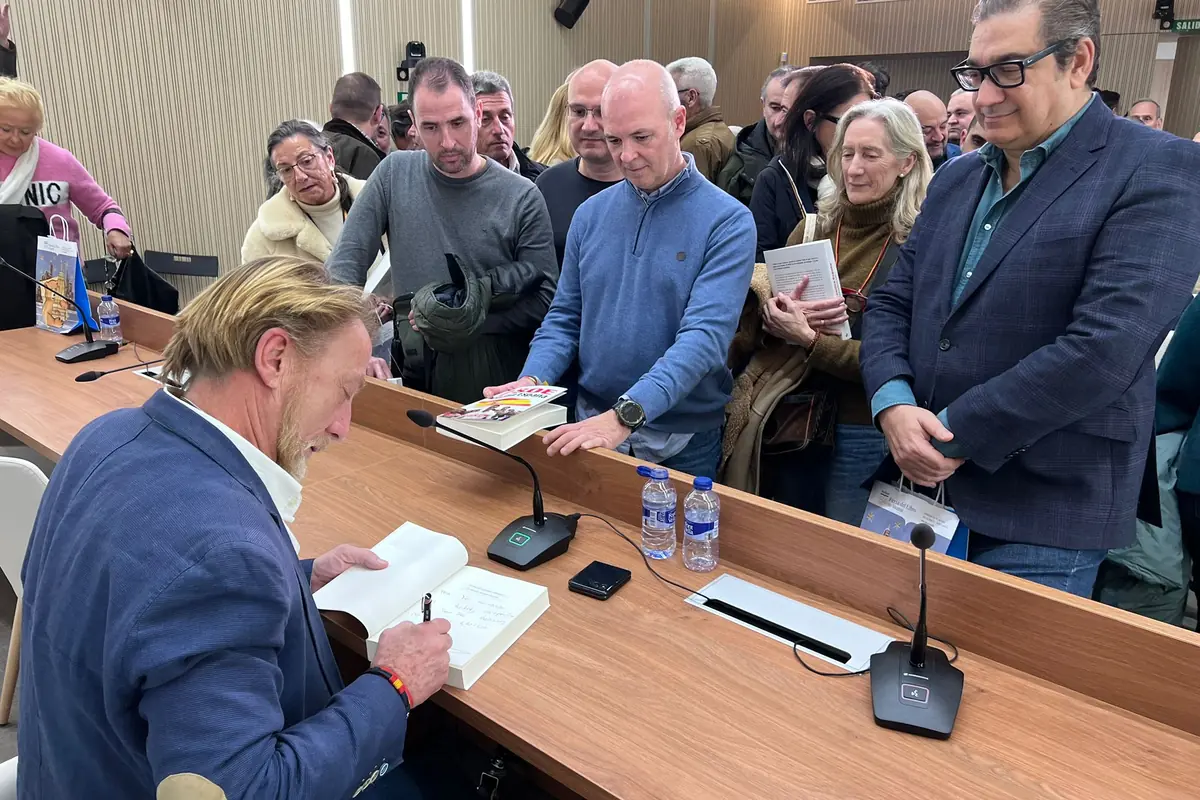 Un hombre firma un libro mientras varias personas esperan en fila con ejemplares en la mano en un evento en un salón iluminado. Un hombre firma un libro mientras varias personas esperan en fila con ejemplares en la mano en un evento en un salón iluminado.
