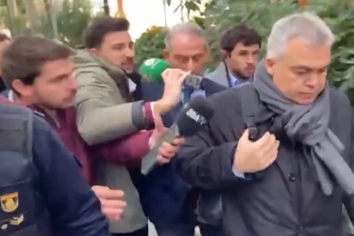 Grupo de periodistas rodea a un hombre mayor con abrigo y bufanda gris mientras le acercan micrófonos y una grabadora en la calle