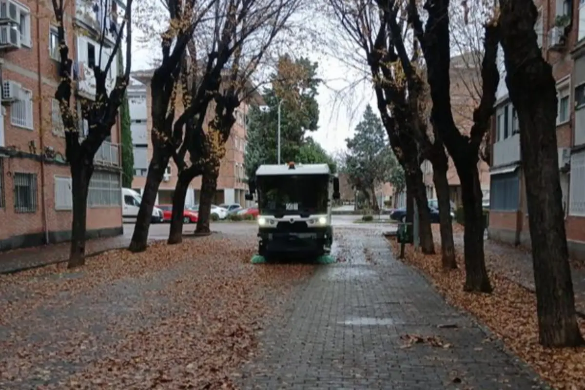 Barredora municipal limpiando una calle arbolada cubierta de hojas secas entre edificios de viviendas