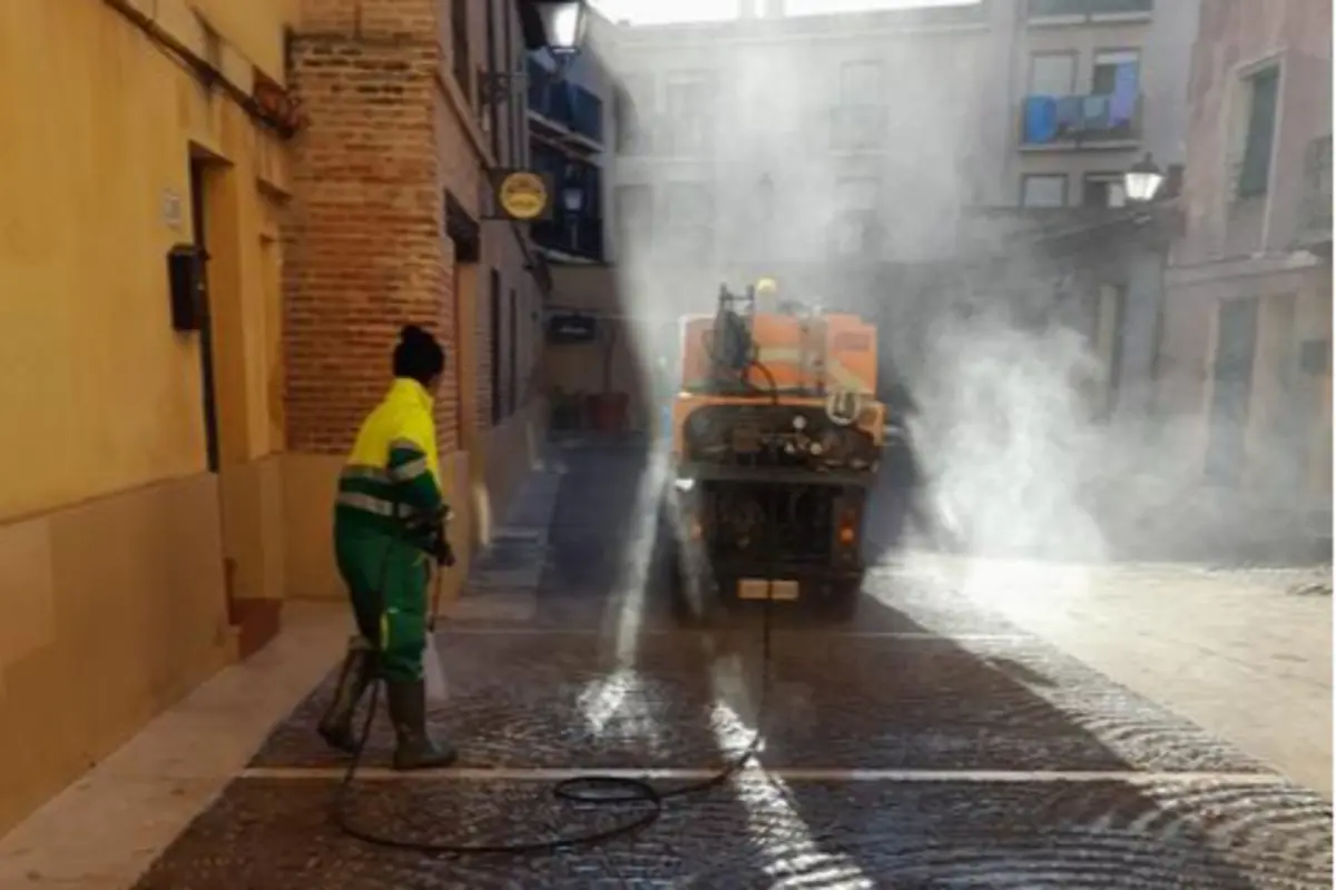 Trabajador de limpieza viaria con manguera a presión desinfectando una calle estrecha junto a un camión cisterna que expulsa vapor