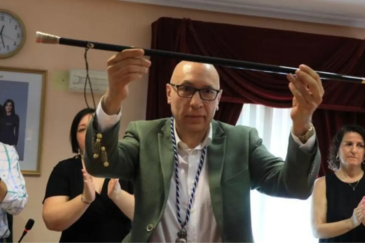 Hombre con gafas y chaqueta verde levantando un bastón ceremonial en una sala mientras varias personas a su alrededor aplauden