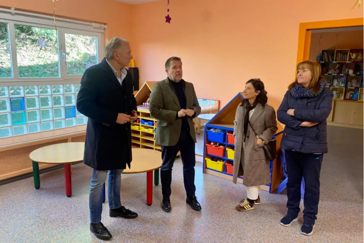 Cuatro personas adultas conversan en el interior de un aula infantil colorida con mesas redondas y estanterías de juguetes
