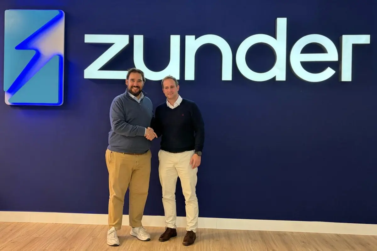 Dos hombres se dan la mano frente a una pared azul con el logo y el nombre de la empresa Zunder