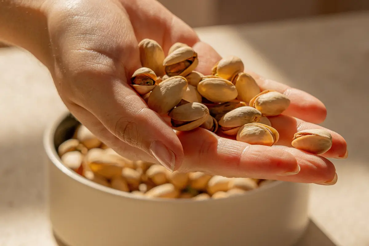 Mano sosteniendo pistachos sobre un cuenco lleno de frutos secos