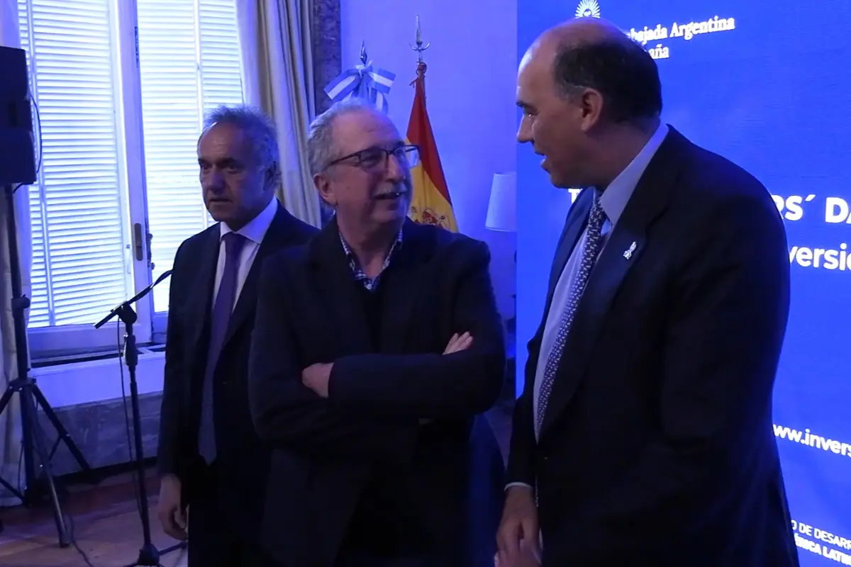 Tres hombres con traje conversan en un evento formal en interiores con banderas de Argentina y España al fondo y una pantalla azul con texto institucional Tres hombres con traje conversan en un evento formal en interiores con banderas de Argentina y España al fondo y una pantalla azul con texto institucional