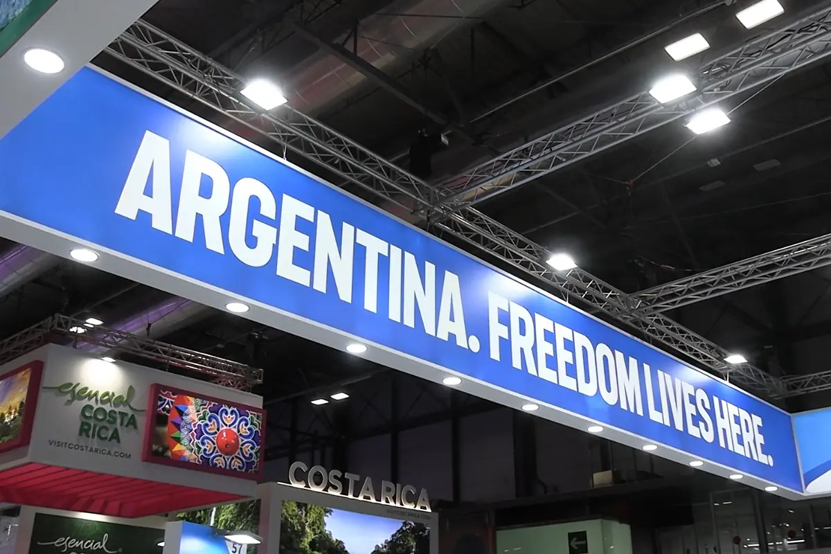 Cartel azul en una feria internacional con el texto ARGENTINA FREEDOM LIVES HERE sobre stands de distintos países Cartel azul en una feria internacional con el texto ARGENTINA FREEDOM LIVES HERE sobre stands de distintos países