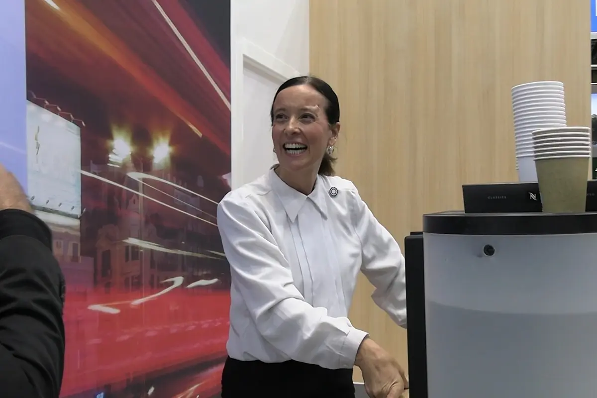 Mujer sonriente con camisa blanca atendiendo una máquina de café en un mostrador con vasos desechables apilados Mujer sonriente con camisa blanca atendiendo una máquina de café en un mostrador con vasos desechables apilados