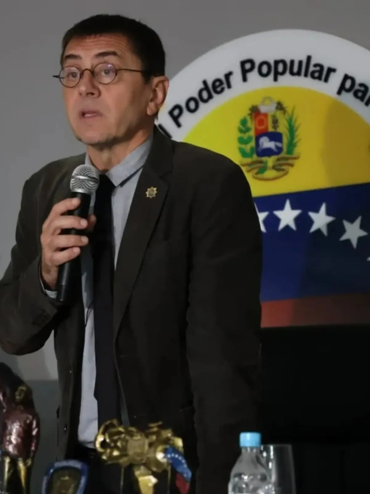 Hombre con gafas y traje oscuro hablando por micrófono frente a un emblema oficial de Venezuela Hombre con gafas y traje oscuro hablando por micrófono frente a un emblema oficial de Venezuela
