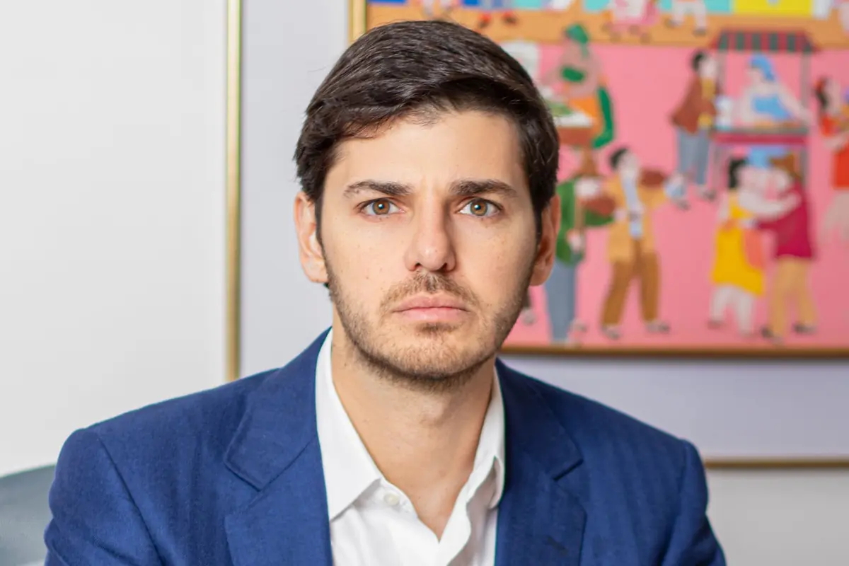 Retrato de un hombre joven con saco azul y camisa blanca mirando a la cámara con expresión seria frente a un cuadro colorido desenfocado de fondo