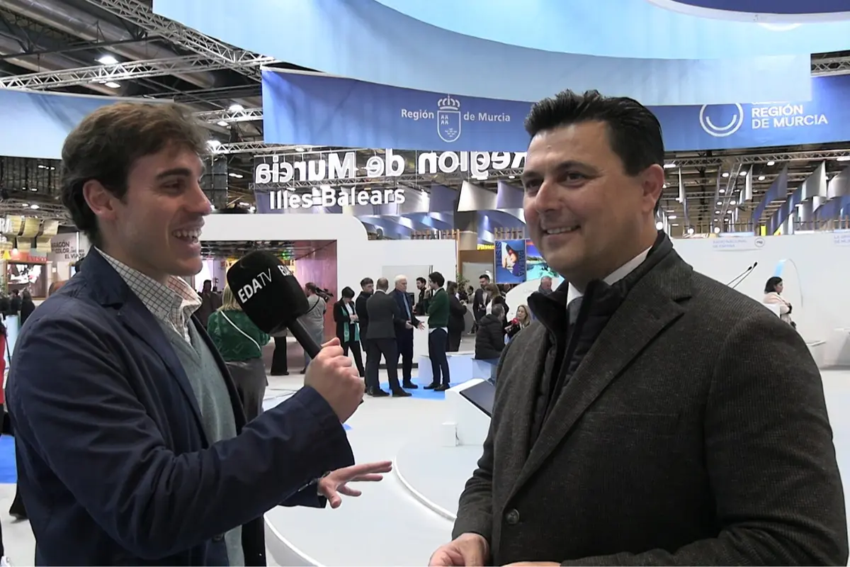 Periodista entrevistando a un hombre en un stand de la Región de Murcia durante una feria, con un micrófono de EDA TV y gente conversando al fondo Periodista entrevistando a un hombre en un stand de la Región de Murcia durante una feria, con un micrófono de EDA TV y gente conversando al fondo