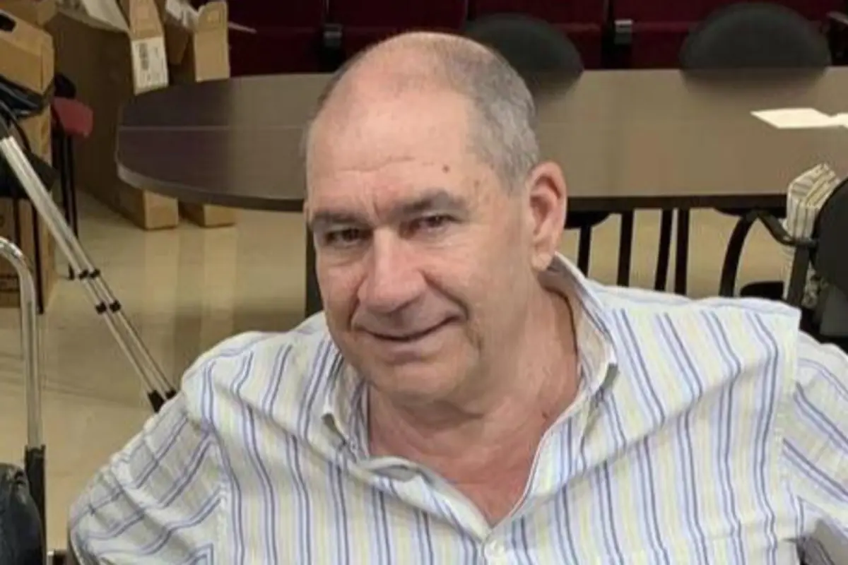 Hombre mayor calvo con camisa de rayas claras sonriendo mientras está sentado en una sala con mesas y sillas de fondo