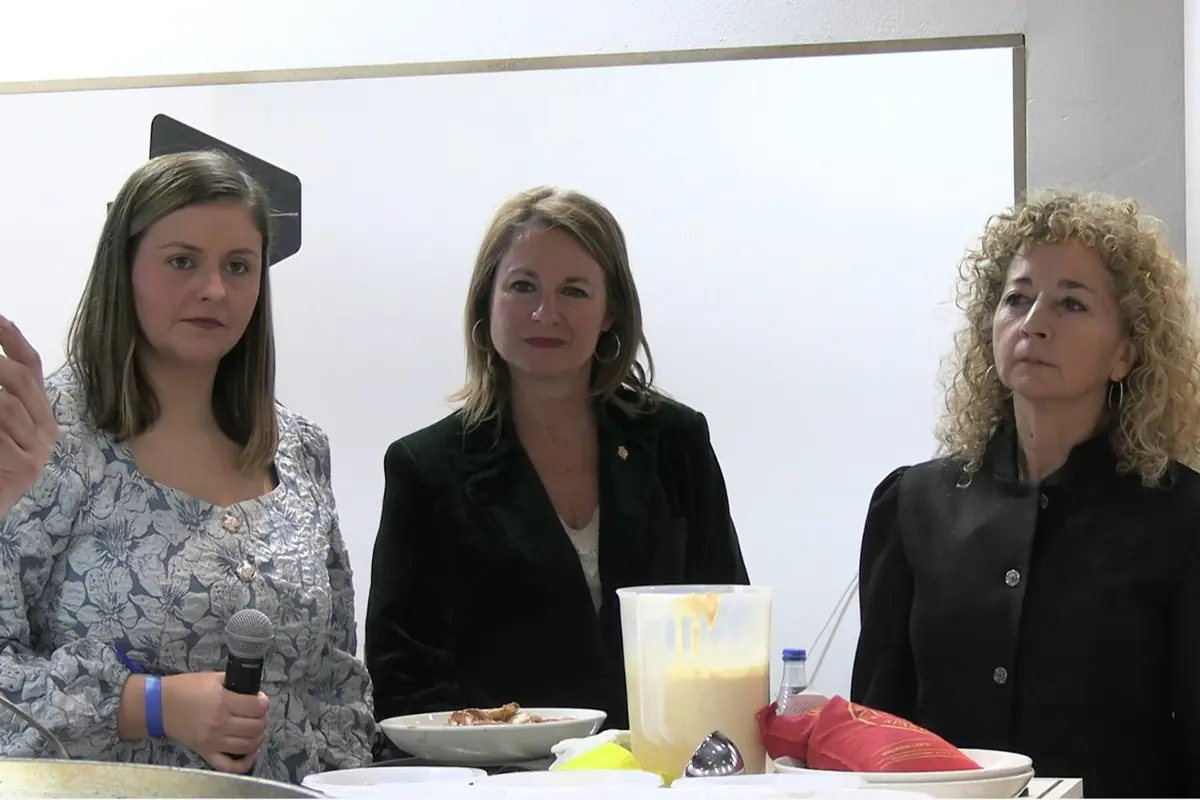Tres mujeres de pie tras una mesa con platos y un recipiente de comida en un entorno interior tipo demostración culinaria