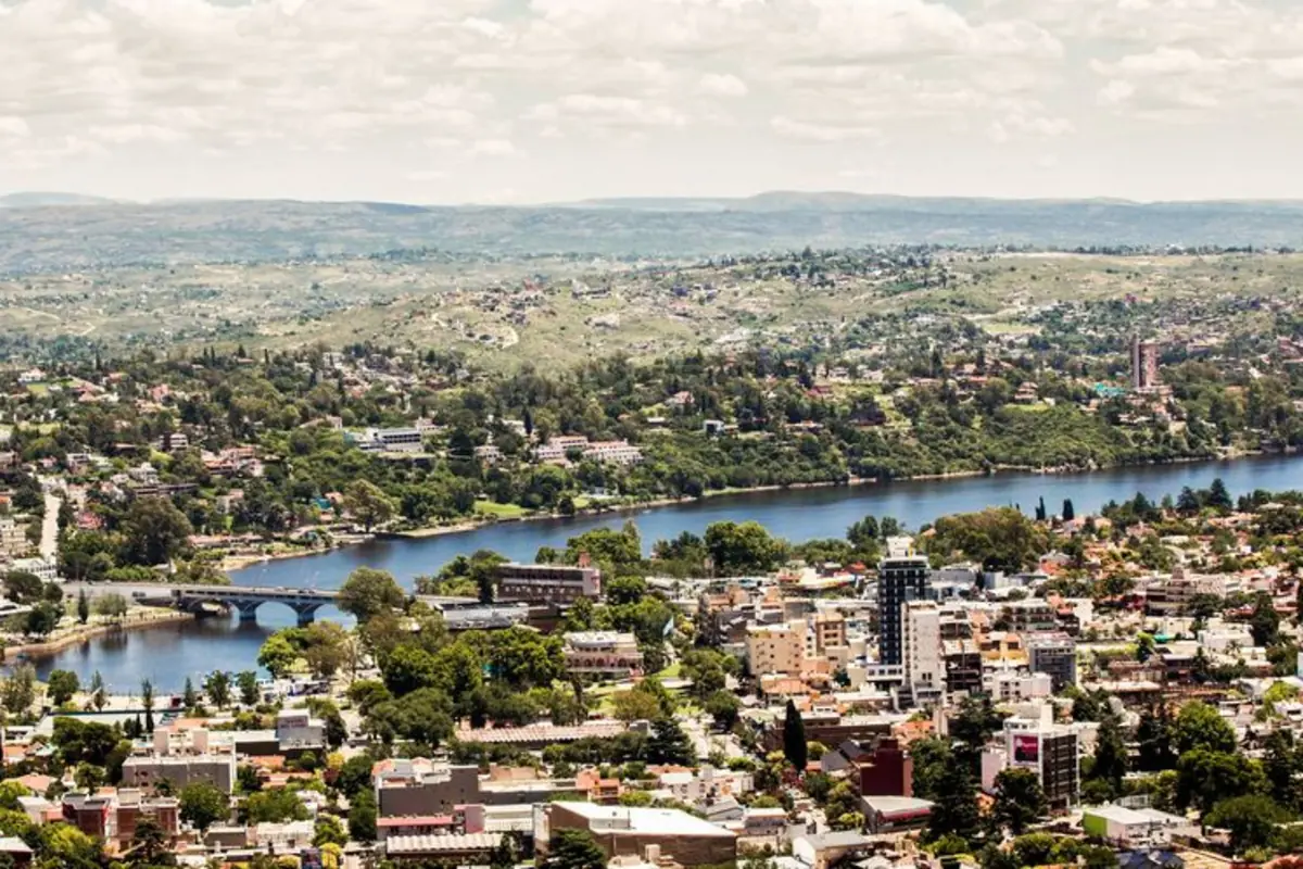 Vista panorámica de una ciudad con edificios bajos y abundante vegetación junto a un río ancho rodeado de colinas verdes bajo un cielo parcialmente nublado