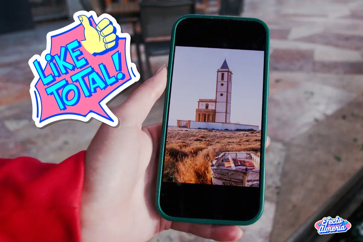 Mano sosteniendo un móvil que muestra la foto de una iglesia junto al mar con un bocadillo de cómic que dice LIKE TOTAL en colores llamativos