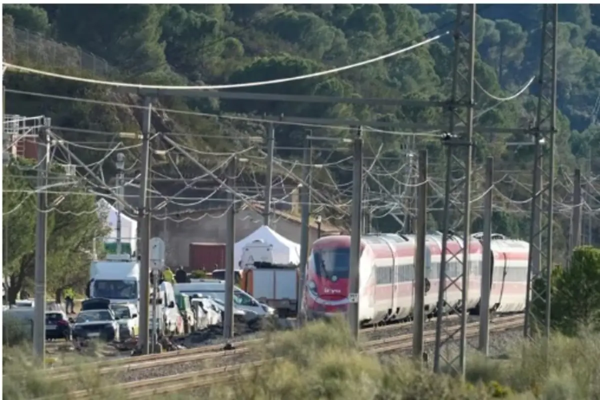 Tren de cercanías rojo y blanco circulando por una vía electrificada entre postes y cables en una zona rural con árboles y varios vehículos de servicio estacionados junto a la vía