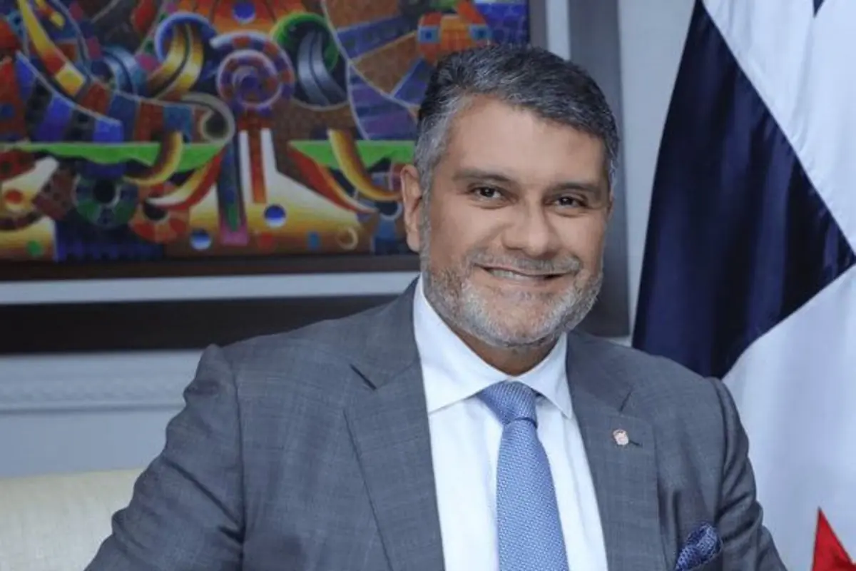 Hombre de mediana edad con barba canosa y traje gris sonriendo frente a una pintura colorida y una bandera en un entorno de oficina