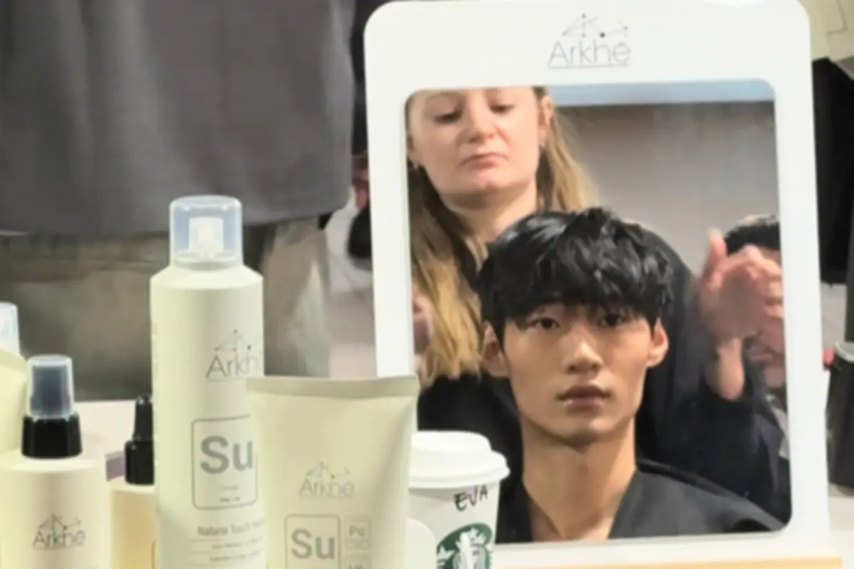 Modelo de cabello sentado frente a un espejo mientras una estilista trabaja detrás de él, con productos de peluquería y un vaso de café sobre la mesa