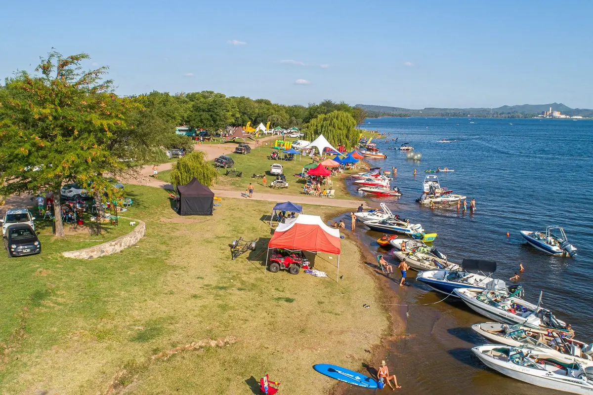 Vista aérea de una playa de lago con muchas lanchas amarradas en la orilla carpas de colores autos estacionados y personas disfrutando del agua y del día soleado