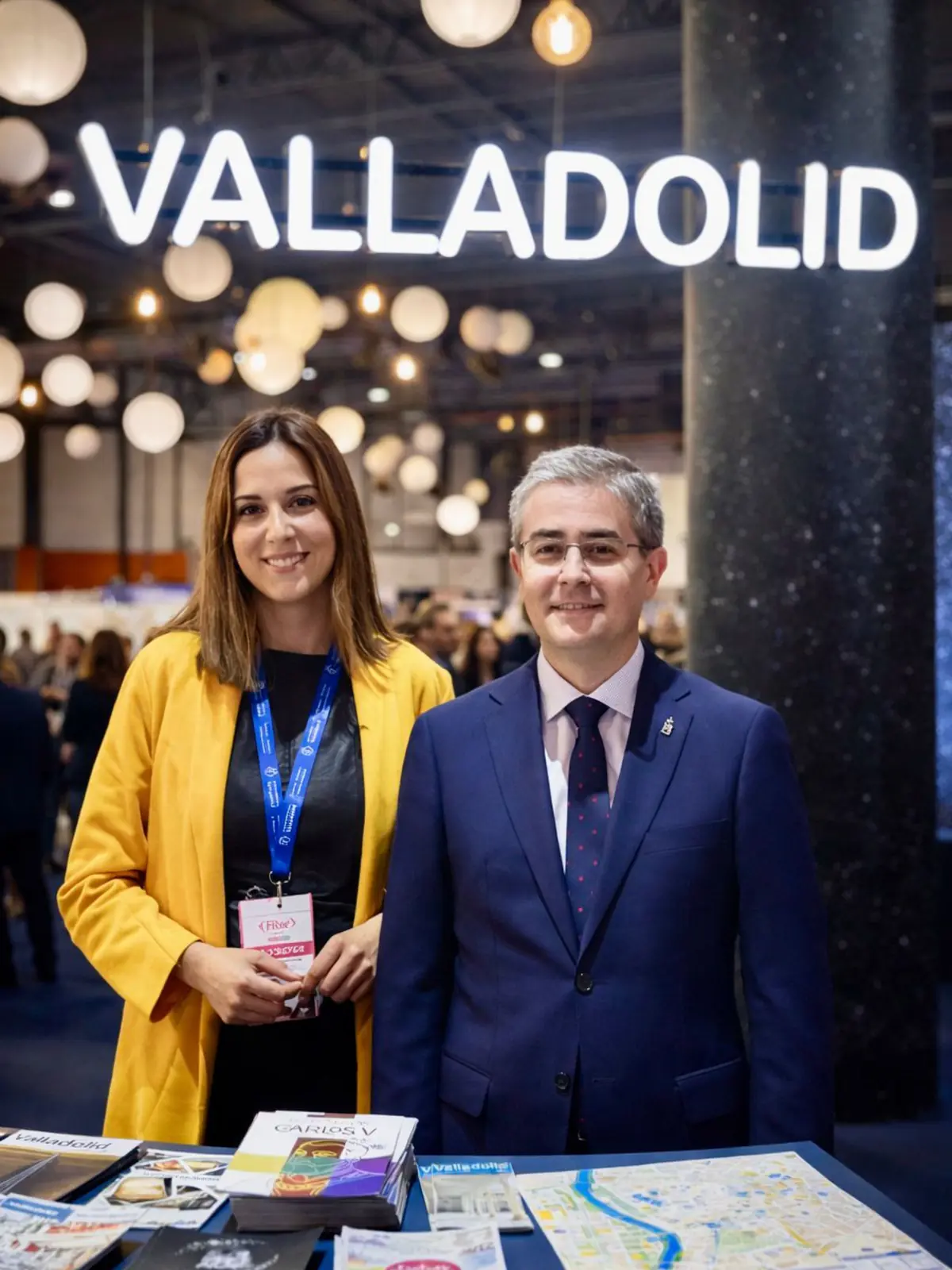 Dos personas posan sonrientes en un stand promocional de Valladolid con folletos y mapas turísticos sobre la mesa y un gran letrero luminoso de la ciudad al fondo Dos personas posan sonrientes en un stand promocional de Valladolid con folletos y mapas turísticos sobre la mesa y un gran letrero luminoso de la ciudad al fondo