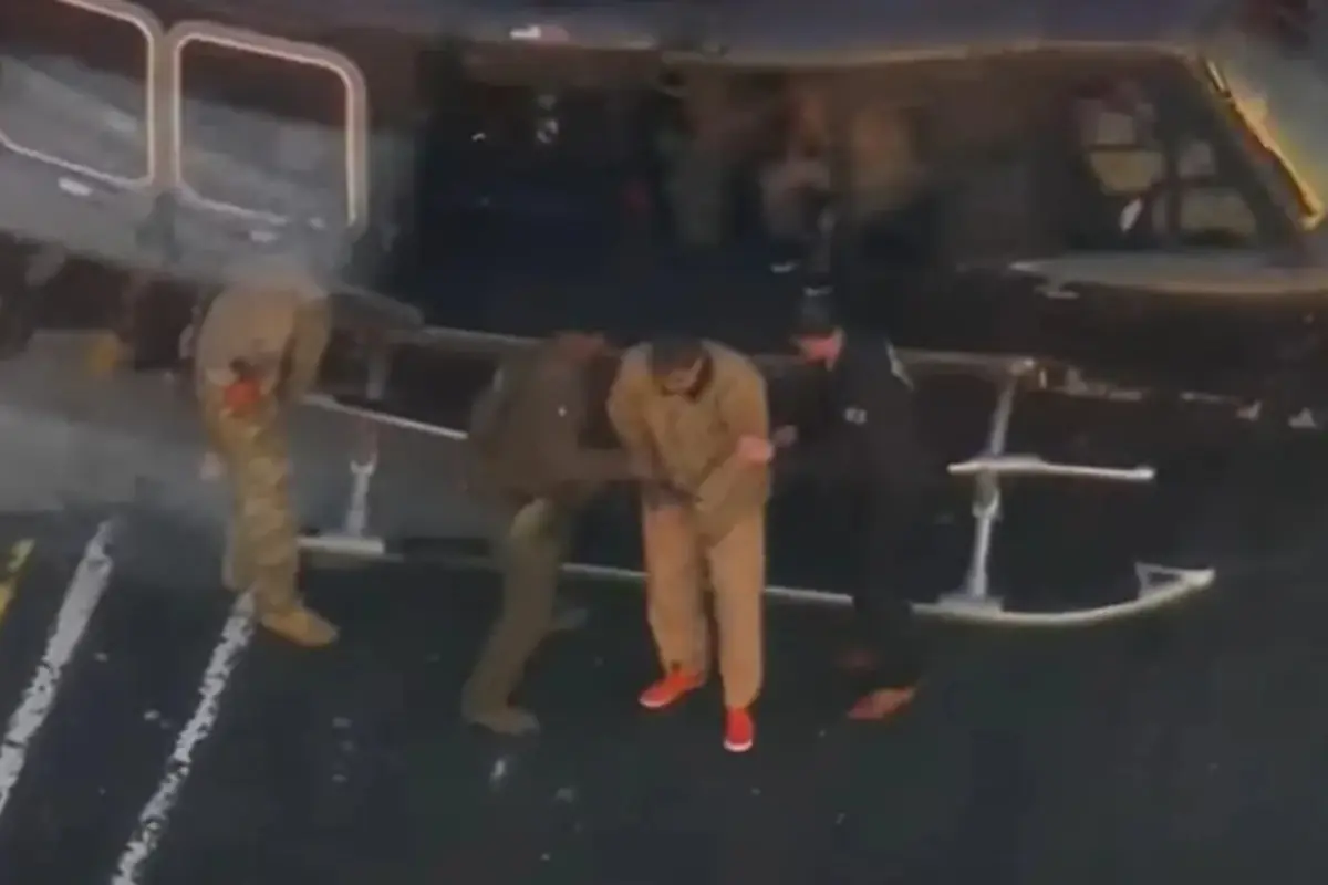 Tres agentes escoltan a un hombre esposado con uniforme beige y tenis naranjas al bajar de un helicóptero negro sobre una pista marcada con líneas blancas Tres agentes escoltan a un hombre esposado con uniforme beige y tenis naranjas al bajar de un helicóptero negro sobre una pista marcada con líneas blancas