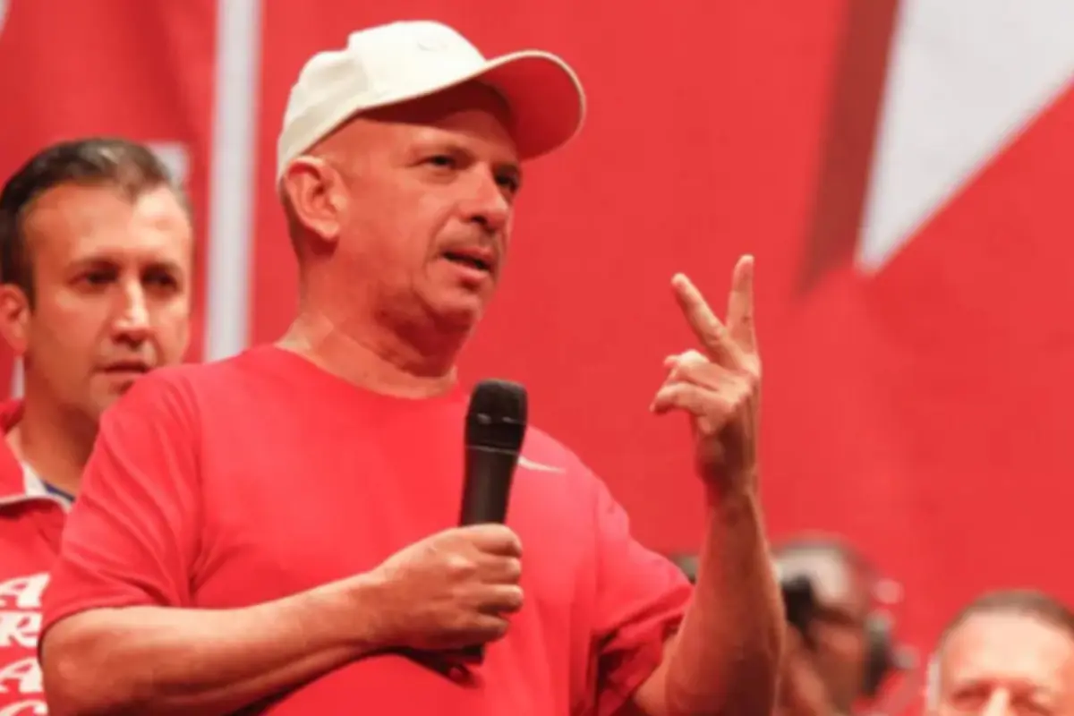 Hombre con gorra blanca y camiseta roja hablando por micrófono y levantando dos dedos frente a un fondo rojo