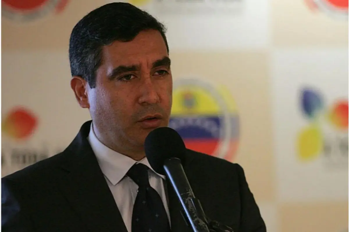 Hombre de traje oscuro hablando frente a un micrófono en un evento oficial con logotipos desenfocados de fondo