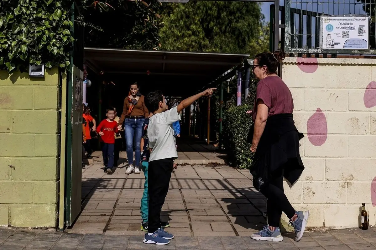 Niño señalando hacia la entrada de una escuela mientras habla con una mujer adulta y otros niños salen al fondo Niño señalando hacia la entrada de una escuela mientras habla con una mujer adulta y otros niños salen al fondo
