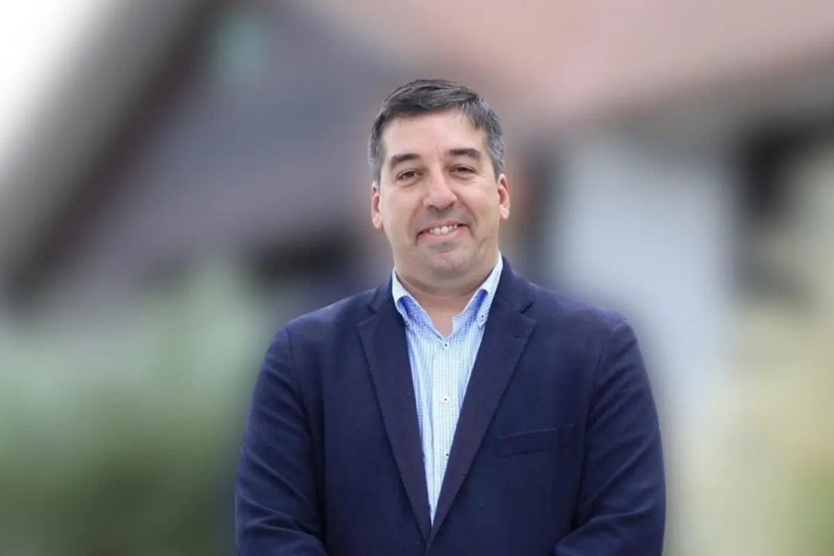 Hombre de mediana edad con saco azul marino y camisa clara sonriendo frente a un fondo urbano desenfocado