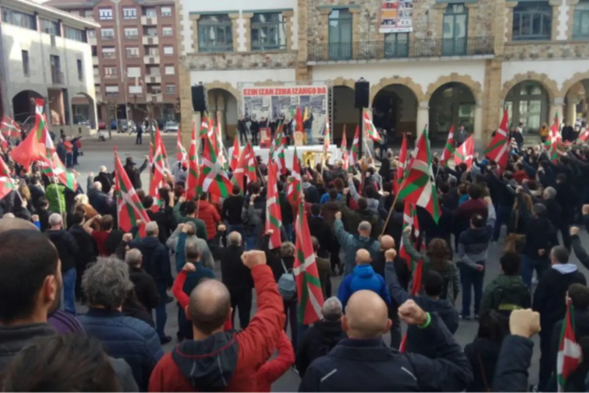 Manifestación multitudinaria en una plaza con numerosas personas levantando el puño y ondeando banderas del País Vasco frente a un escenario con pancartas y altavoces Manifestación multitudinaria en una plaza con numerosas personas levantando el puño y ondeando banderas del País Vasco frente a un escenario con pancartas y altavoces