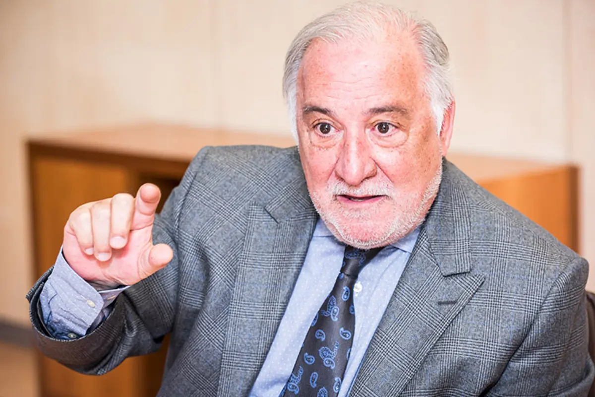 Hombre mayor con traje gris gesticulando con la mano mientras habla en un entorno de oficina