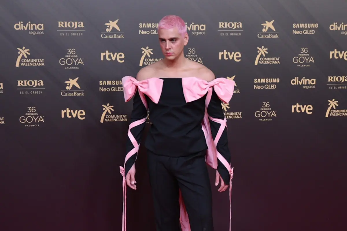 Persona con el pelo rosa posa en una alfombra roja vistiendo un conjunto negro con grandes lazos y tiras rosas en los hombros y brazos frente a un fondo con logotipos dorados de patrocinadores