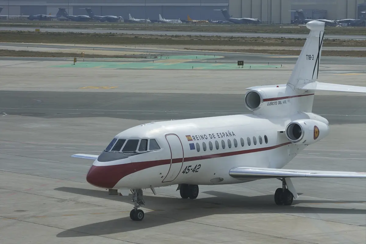 Avión oficial del Reino de España estacionado en la pista de un aeropuerto con hangares y otros aviones al fondo