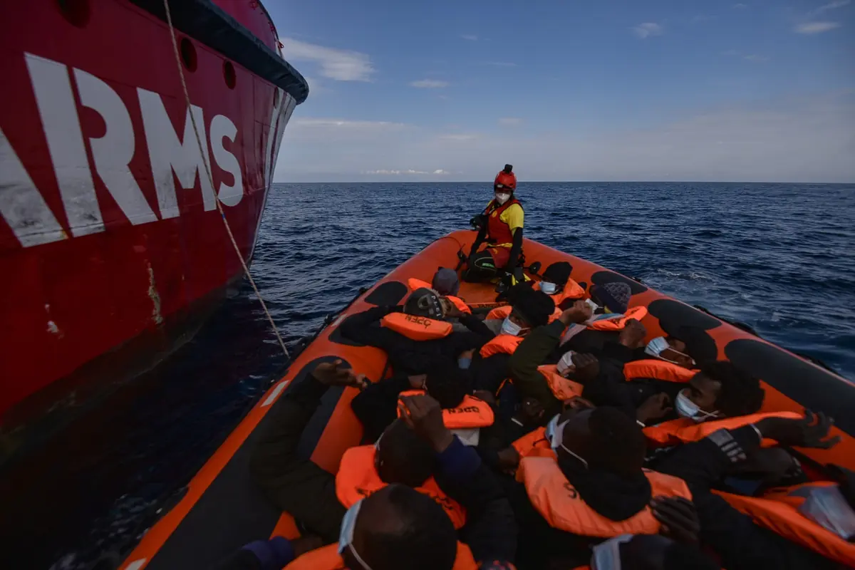 Personas con chalecos salvavidas naranjas en una lancha neumática abarrotada se acercan a un gran barco de rescate rojo en mar abierto