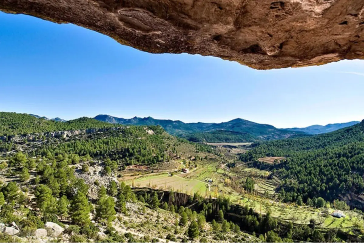 Vista panorámica de un valle verde con campos de cultivo y colinas boscosas bajo un cielo azul despejado enmarcado por la roca de una cueva en primer plano