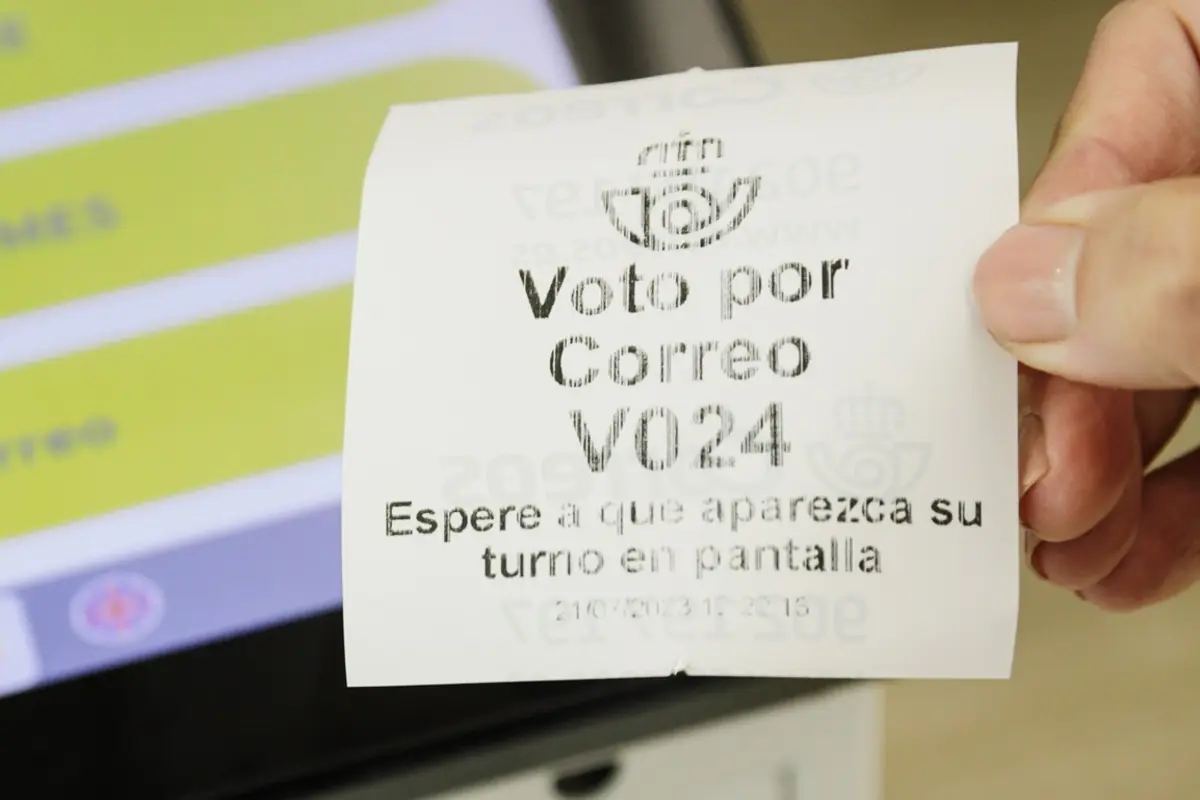 Mano sosteniendo un ticket de turno para voto por correo con el número V024 frente a una pantalla táctil