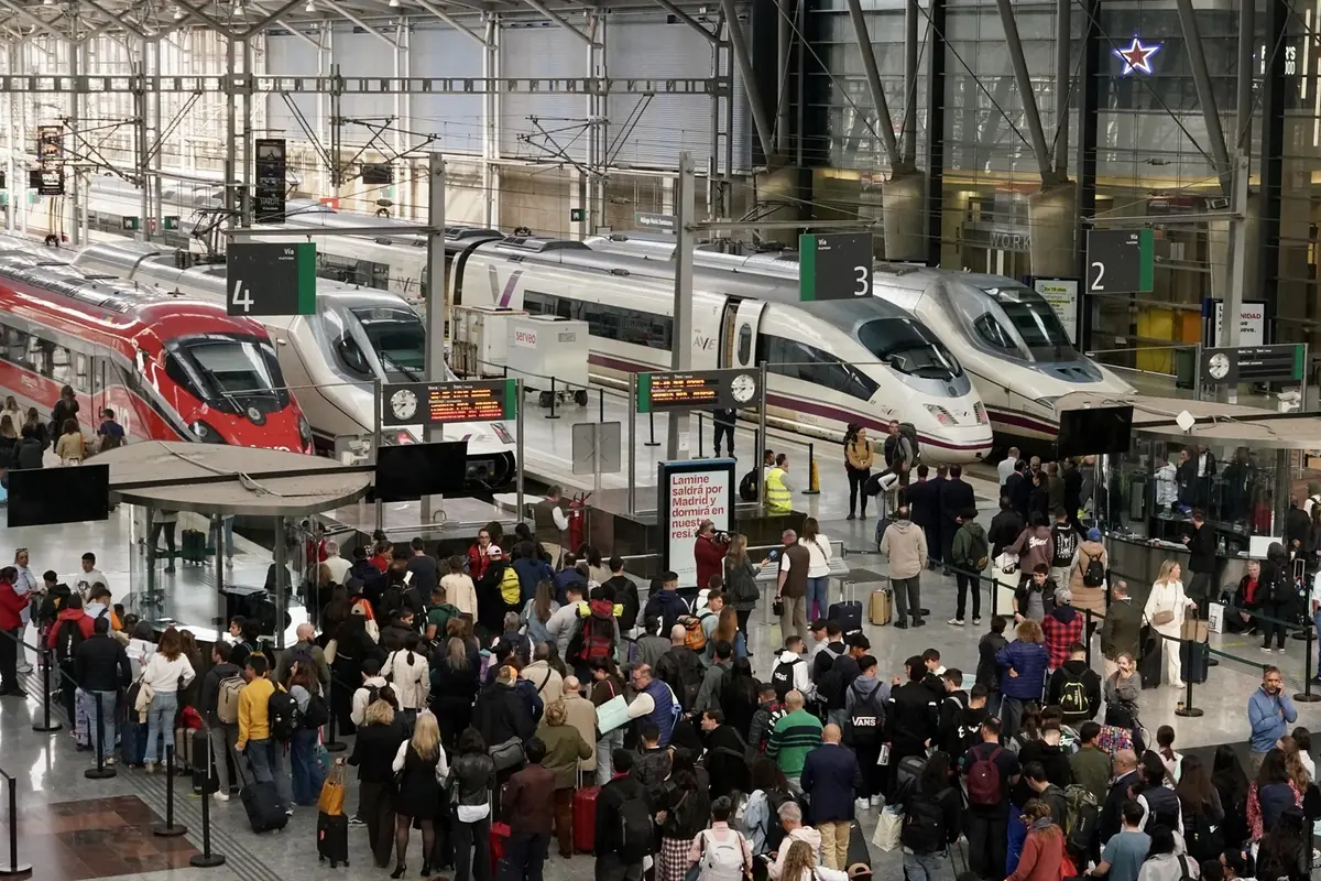 Andén de estación de tren de alta velocidad con varios trenes AVE y un tren rojo mientras una gran multitud de pasajeros hace fila con su equipaje