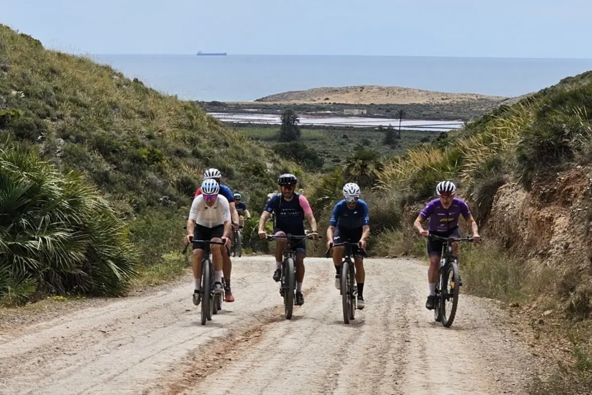 Grupo de ciclistas pedaleando por un camino de tierra entre colinas verdes con el mar al fondo