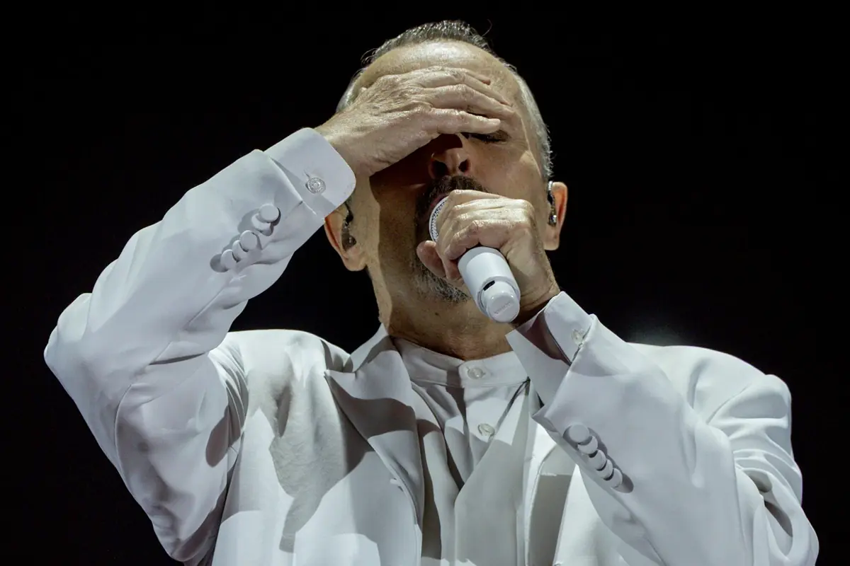 Cantante con traje blanco sosteniendo un micrófono mientras se cubre los ojos con una mano en el escenario