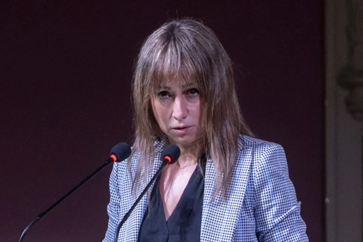 Mujer de mediana edad con chaqueta de cuadros hablando seria frente a dos micrófonos en un auditorio