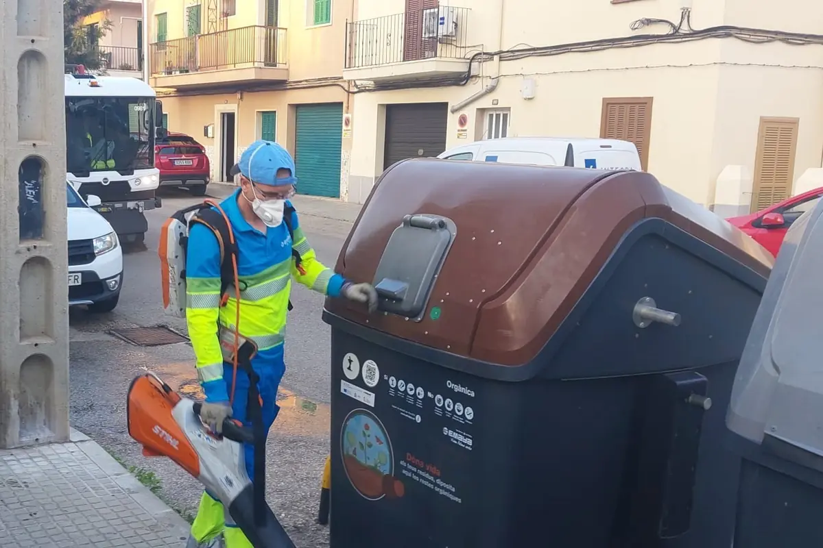 Trabajador de limpieza con mascarilla y sopladora usando un contenedor de residuos orgánicos en una calle urbana