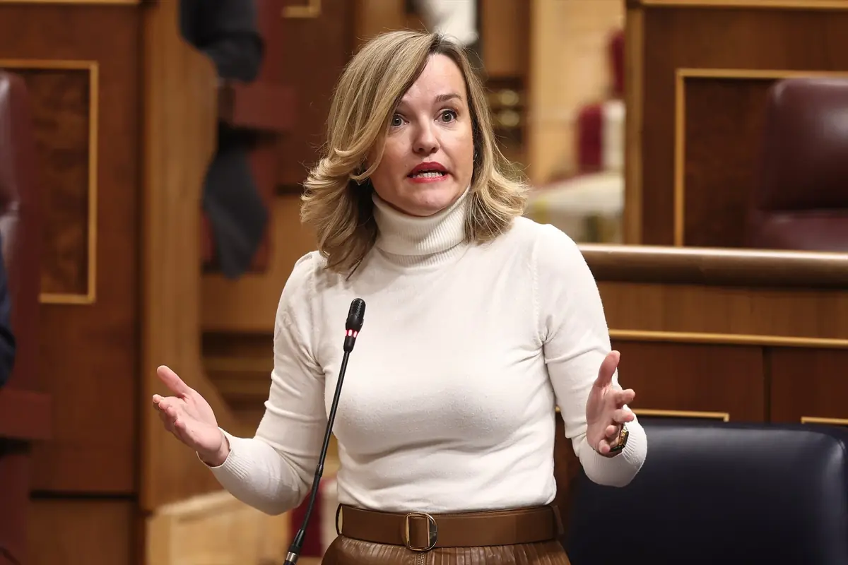 Mujer rubia con jersey blanco hablando gesticulando frente a un micrófono en un parlamento