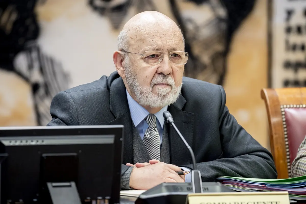 Hombre mayor con barba canosa y traje formal sentado en una mesa de reuniones frente a un micrófono y documentos