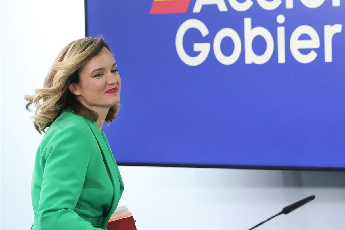 Mujer con chaqueta verde y carpeta en la mano caminando frente a una pantalla azul con texto parcial de un mensaje institucional Mujer con chaqueta verde y carpeta en la mano caminando frente a una pantalla azul con texto parcial de un mensaje institucional
